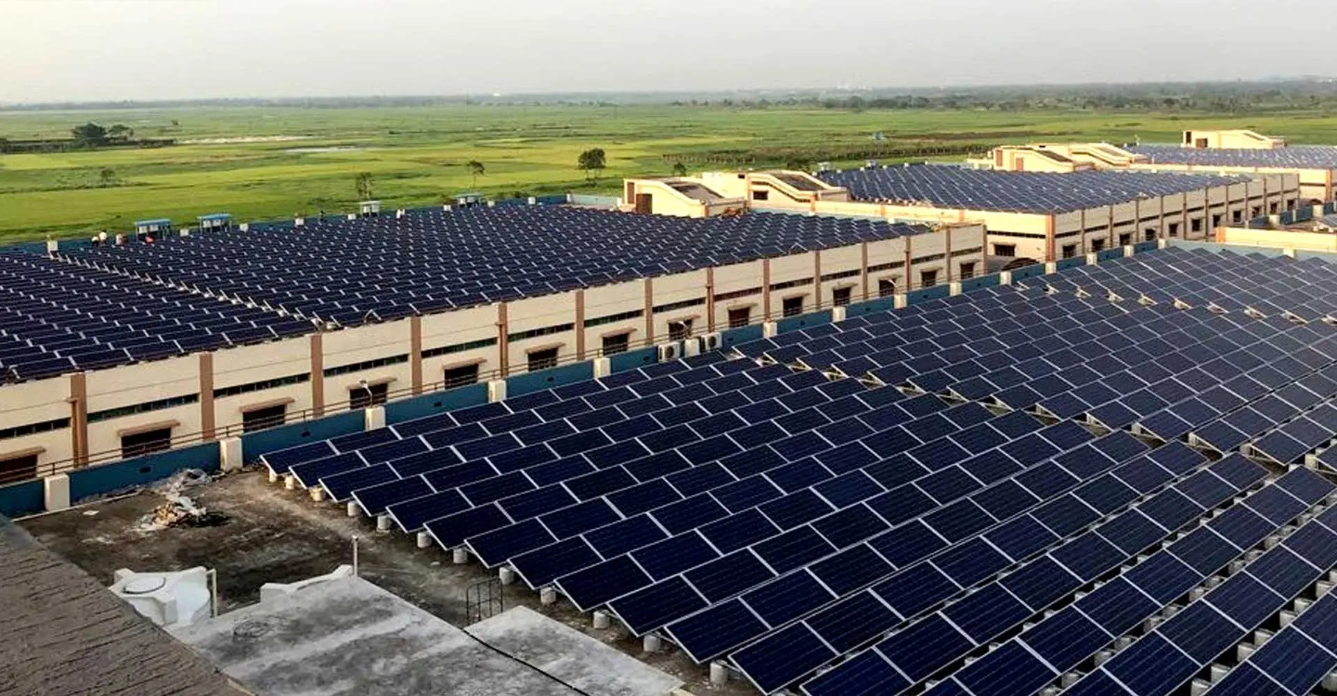 A large rooftop solar power plant across several factory buildings, with green fields beyond